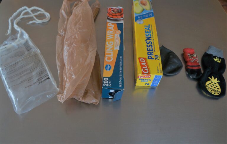dog broken leg cast protection tips -- iv bag, grocery bag, plastic wrap, press & seal, various dog booties -- sock kind and one galoshes kind laid out left to right on a stainless steel table